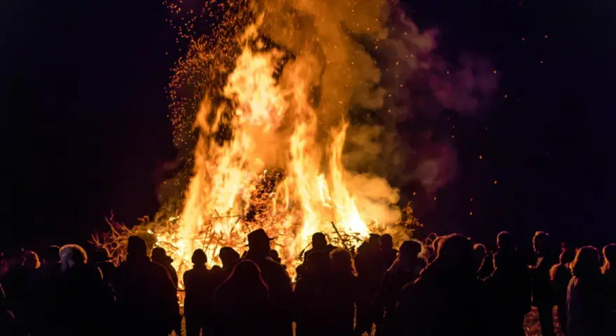 Menschen versammeln sich um ein helles Osterfeuer. Die Flammen lodern hoch in den Nachthimmel, Funken fliegen. Das Osterfeuer symbolisiert die christliche Bedeutung von Licht und Auferstehung.