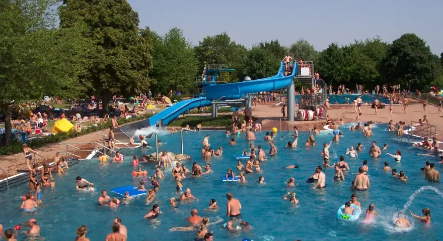 Ein belebtes Freibad in Goslar mit vielen Menschen, die im Wasser planschen und die blaue Wasserrutsche genießen.