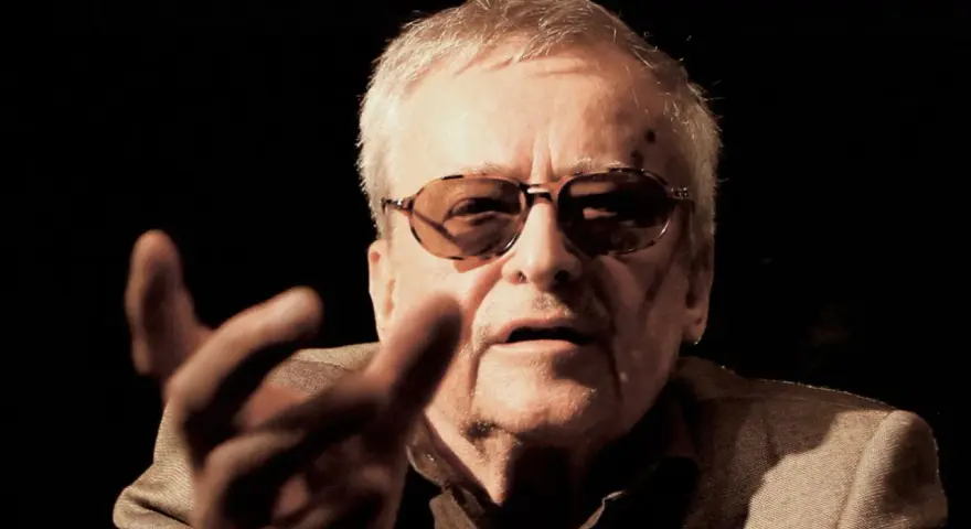 Jerzy Jarocki w okularach, z gestem dłoni, w ciemnym tle.