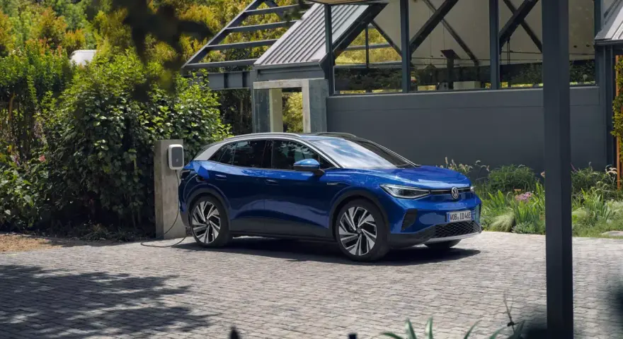 Niebieski Volkswagen ID.4 ładuje się przy domu, otoczony zielenią.