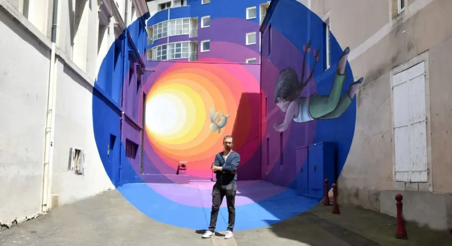 Un artiste street art français pose devant sa fresque colorée d'une fillette et d'un lapin flottant dans un tunnel psychédélique.