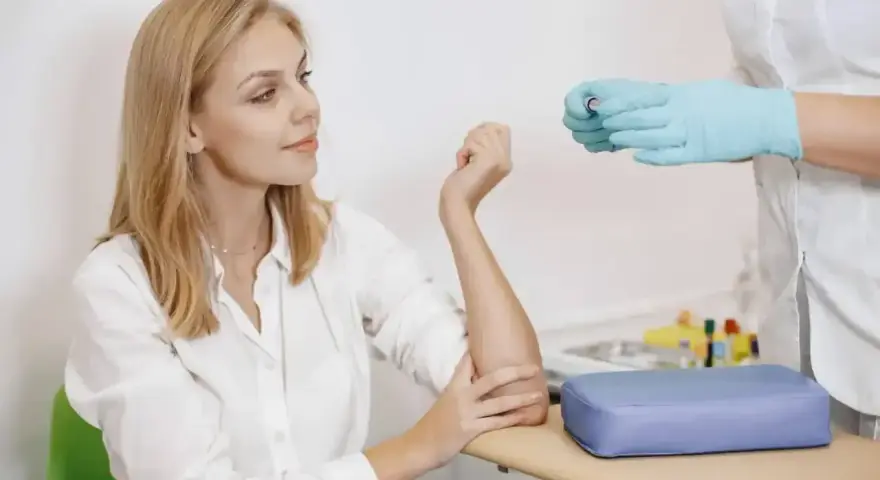 Kobieta w białej koszuli przygotowuje się do pobrania krwi. Pielęgniarka w rękawiczkach trzyma probówkę. To standardowa procedura, gdy badanie wykonuje się na czczo.