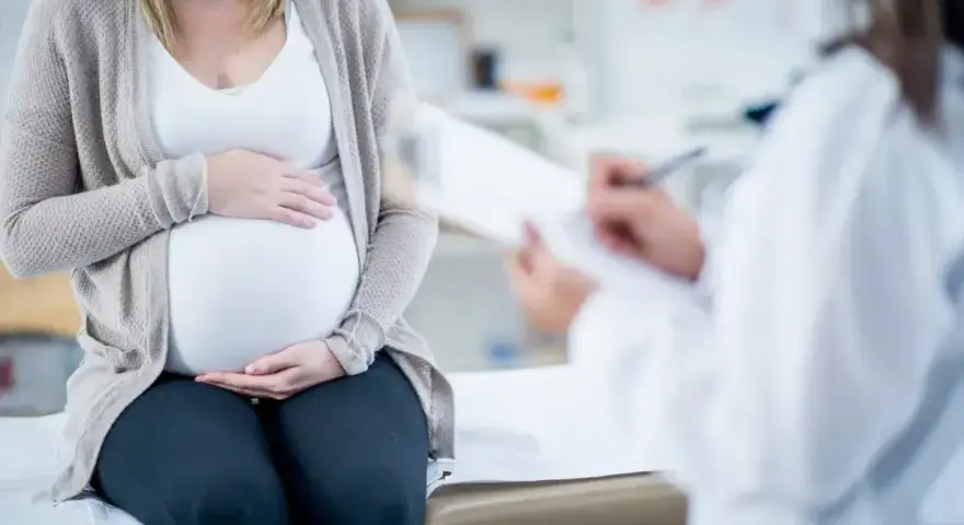 Kobieta w ciąży siedzi na kozetce, trzymając dłonie na brzuchu. Lekarz spisuje dokumenty, prawdopodobnie wystawiając zaświadczenie o ciąży.