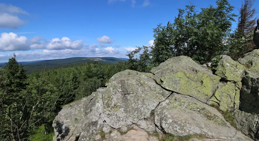 Kamienne formacje na rozdrożu izerskim, porośnięte mchem, z widokiem na gęsty las i błękitne niebo z chmurami.
