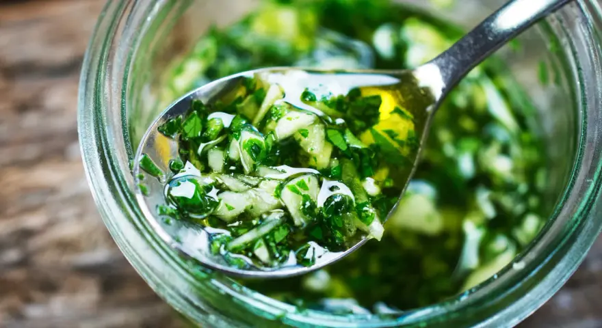 Łyżka z aromatycznym, zielonym sosem chimichurri, pełnym ziół i czosnku, zanurzona w słoiku.