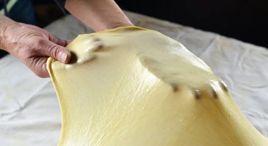 Mani che stendono la pasta matta per una ricetta tradizionale. La pasta è sottile e traslucida, con le dita che creano delle leggere ondulazioni.