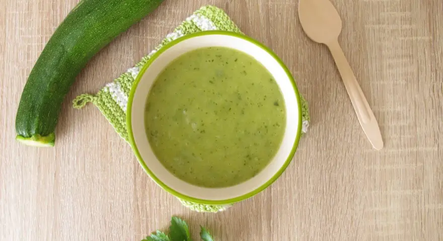 Zucchini Brei Baby: Eine Schüssel grüner Zucchinipüree, daneben eine Zucchini und ein Holzlöffel.