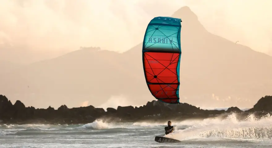 Kitesurfer na desce w akcji, z kolorowym latawcem na tle gór i morza.
