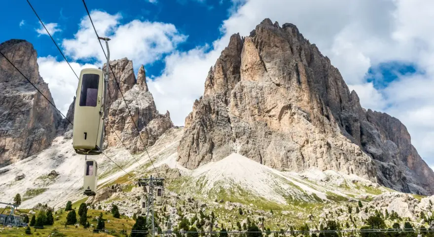 Cabine della funivia salgono verso le imponenti vette del Sassolungo, un anello di roccia maestoso sotto un cielo azzurro con nuvole bianche.