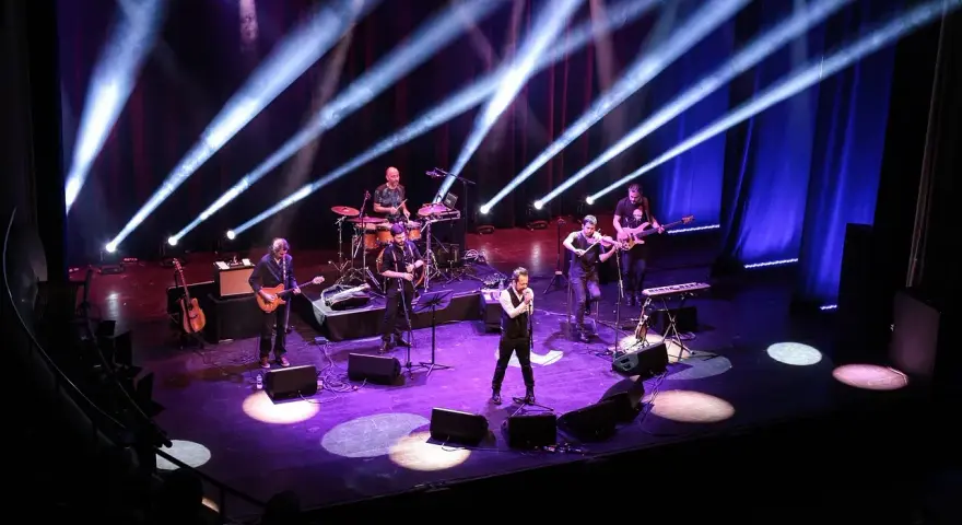 Zespół muzyczny na scenie, oświetlony niebieskimi i fioletowymi światłami. Wokalista na pierwszym planie, gitarzyści, basista, skrzypek i perkusista. Jak liczyć takty w muzyce, można się nauczyć, obserwując ich rytm.
