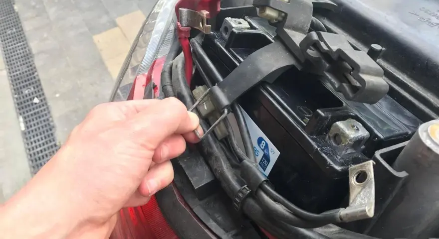 Ręka odkręca mocowanie akumulatora w skuterze. Widać jego umiejscowienie pod siedzeniem.