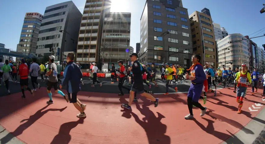 Atleti corrono nella maratona di Tokyo, circondati da grattacieli moderni sotto un sole splendente.