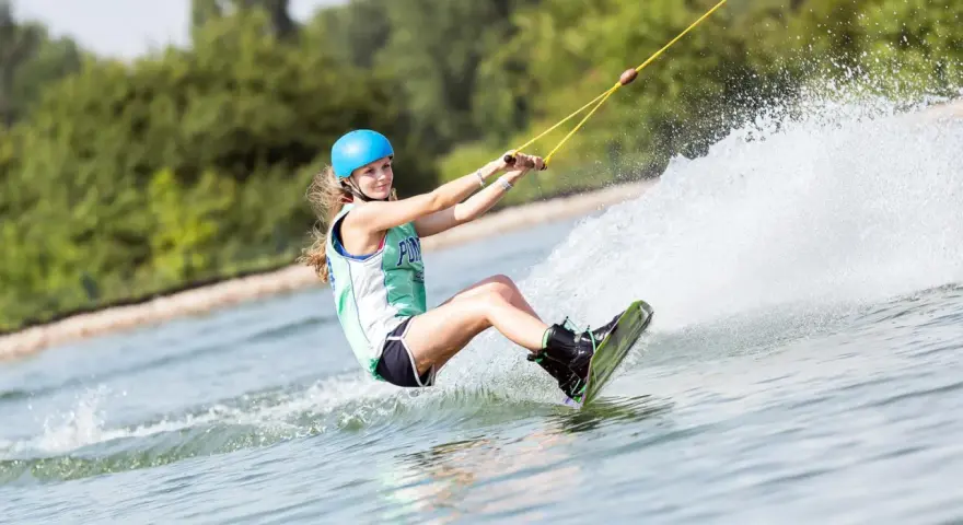 Dziewczyna w kasku i kamizelce pływa na wakeboardzie, tworząc wielki szelment wody.
