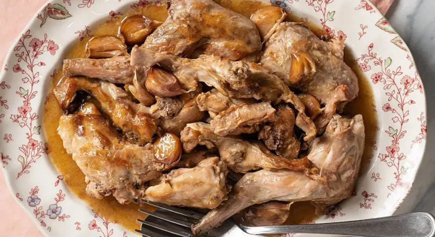 Conejo al ajillo servido en un plato con borde floral, con trozos de carne dorada y dientes de ajo enteros.