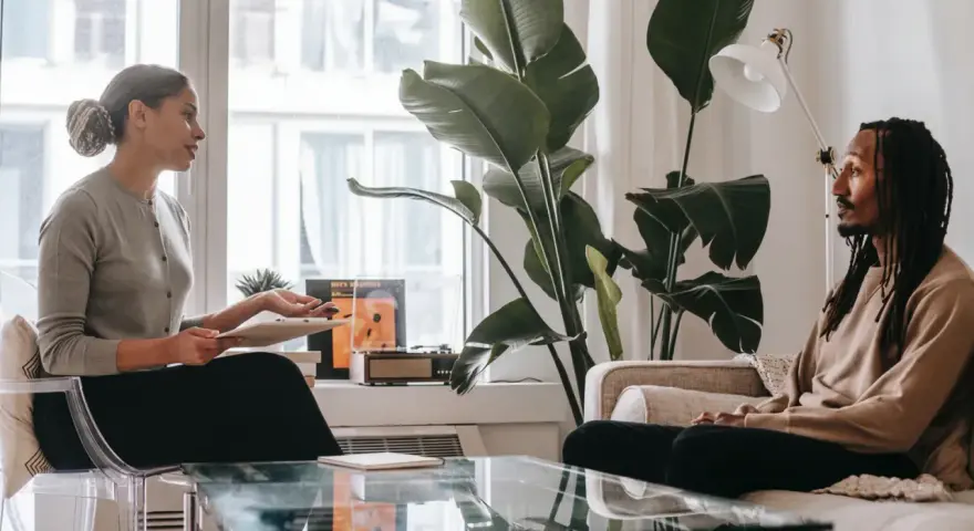 En kvinna och en man sitter mitt emot varandra i ett rum. Kvinnan håller en surfplatta och talar. Mannen lyssnar. Kanske diskuterar de hur mycket kostar terapi.