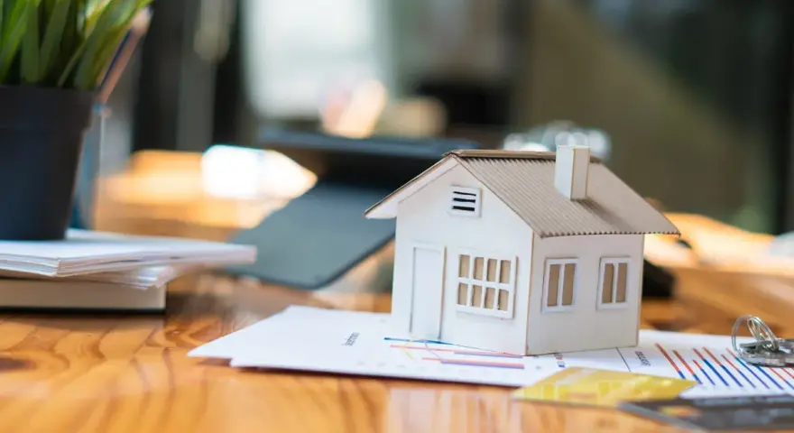 Maqueta de casa sobre gráficos, sugiriendo cómo saber si una casa tiene cargas. Llaves y tarjetas de crédito cerca.