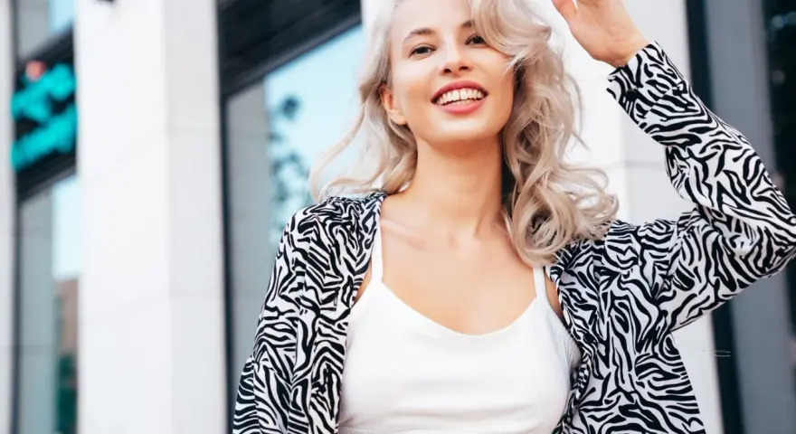 Uśmiechnięta blondynka z falowanymi włosami, efekt farbowania włosów na blond. Wzorzysta marynarka i biały top.