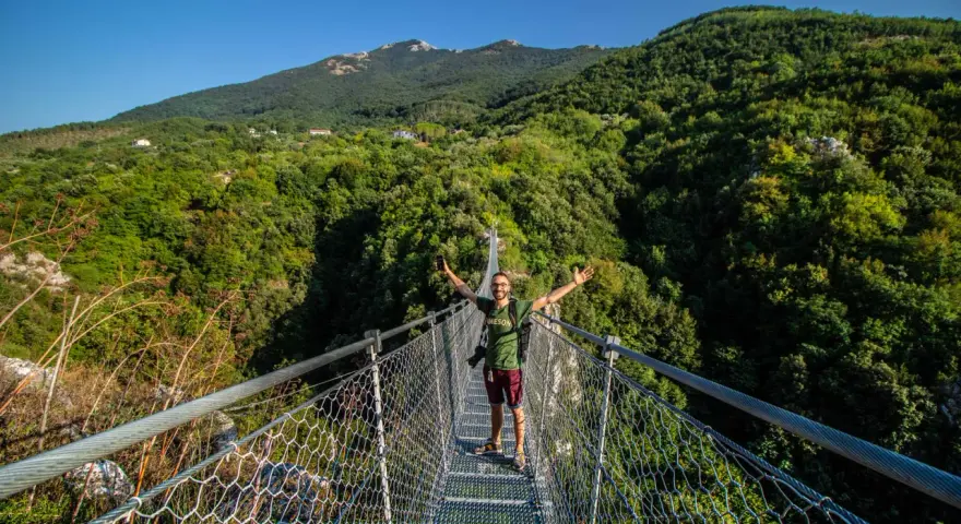 Uomo felice sul laviano ponte tibetano, con montagne verdi e cielo azzurro sullo sfondo.