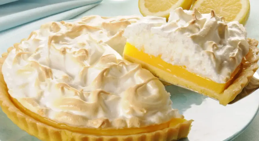 Pyszna tarta cytrynowa z bezą, z kawałkiem na łopatce. Obok leżą połówki cytryn.