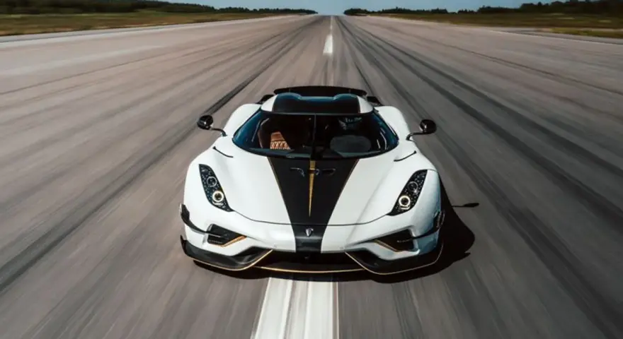 Biały Koenigsegg Regera z czarnym pasem i złotymi akcentami pędzi po pasie startowym, zostawiając za sobą smugę prędkości.