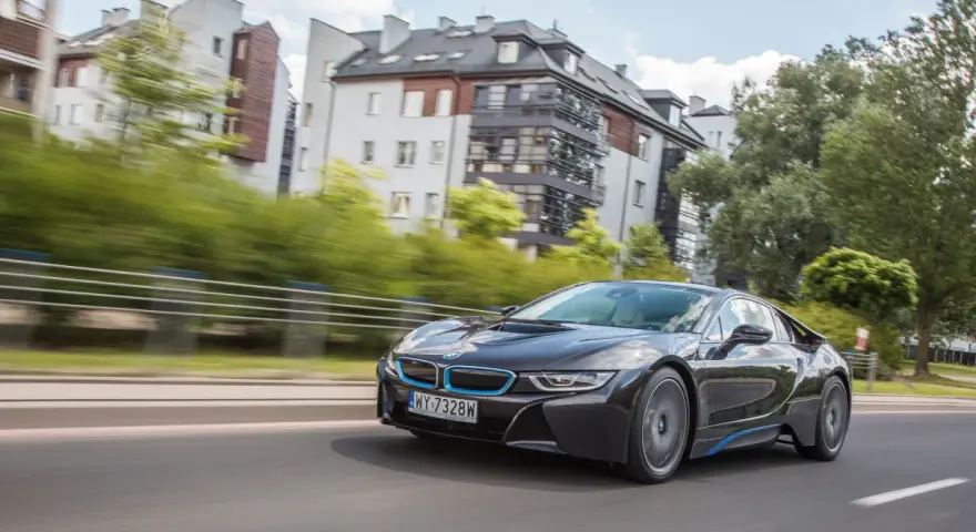 Czarny BMW i8 pędzi ulicą, mijając nowoczesne budynki i zielone drzewa.