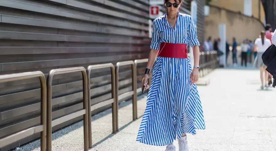 Donna con vestito a righe blu e bianche, cintura rossa e sneakers bianche cammina per strada.