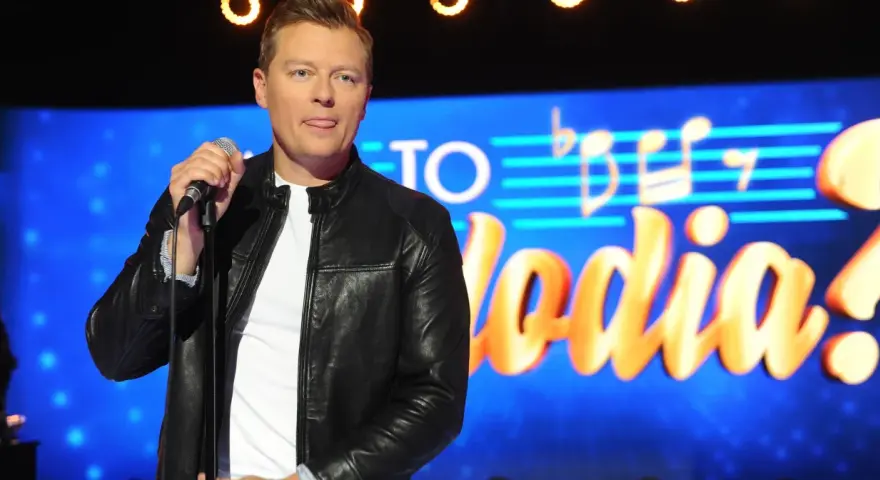 Rafał Brzozowski z mikrofonem na scenie, w tle napis "Jaka to melodia?".