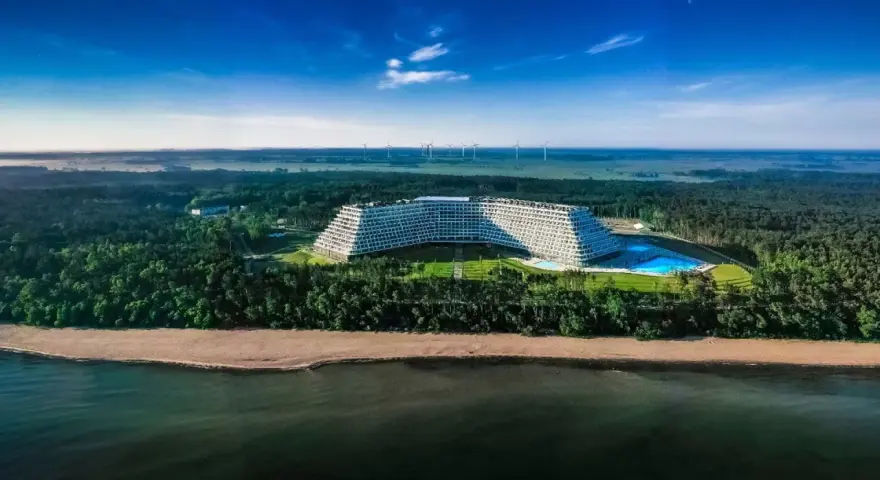 Hotel Gołębiewski w Pobierowie, otoczony lasem, z basenem i widokiem na morze. W oddali widać farmę wiatrową.