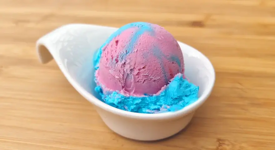Ein Löffel eis mit kaugummi Geschmack, in rosa und blau, serviert in einer weißen Schale auf Holzuntergrund.