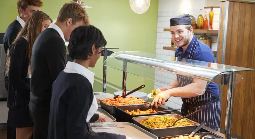 Des élèves font la queue pour le déjeuner à la cantine. Un employé souriant sert des légumes. Un contrat extra restauration est en cours de discussion.