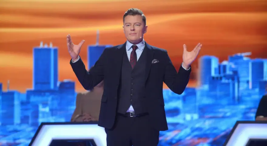 Rafał Brzozowski w garniturze, z rękami uniesionymi na boki, prowadzi program "Jaka to melodia?".