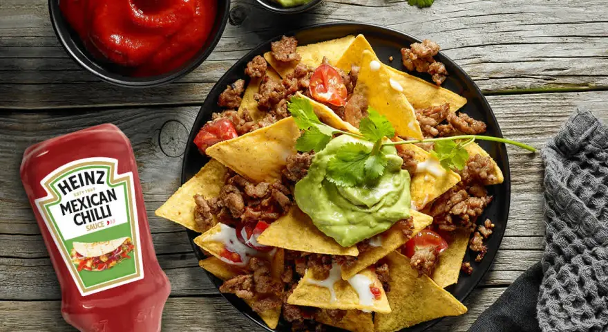Nachos con carne, guacamole y salsa en un plato negro, con botella de Heinz y cuencos de salsa.