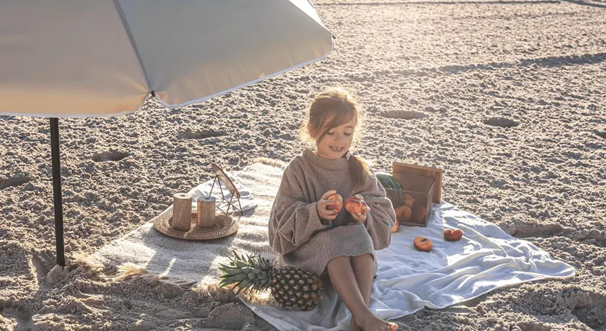 Dziewczynka na plaży pod parasolem, z owocami i ananasem. Idealny zestaw, by wiedzieć, co zabrać na wakacje nad morze z dzieckiem.