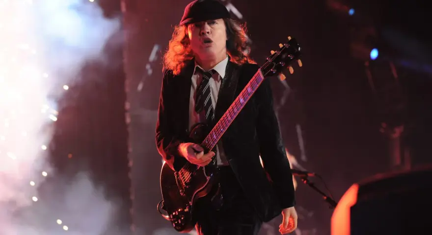 Ein Mann mit Hut und Schlagzeug spielt auf einer ACDC Gitarre, umgeben von Rauch und Lichtern.