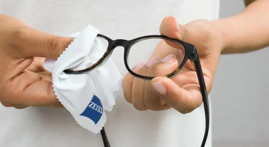 Här putsas glasögon med en mjuk trasa. En person håller i bågarna och putsar linserna med en vit trasa med Zeiss-logga.