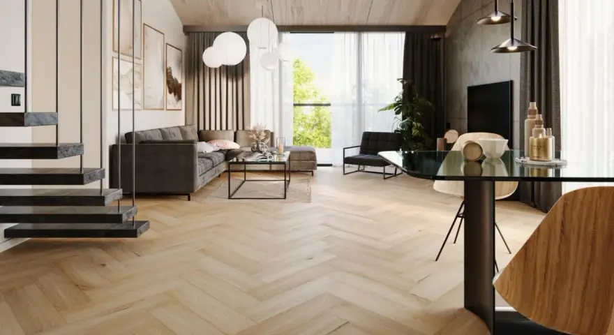Nowoczesny salon z szarą sofą, szklanym stolikiem i jadalnią. Wzrok przyciągają panele bez dylatacji w progu, tworzące jednolitą, drewnianą podłogę w jodełkę.