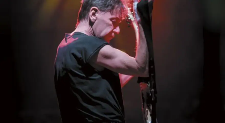 Muzyk z zespołu KSU z gitarą na scenie, w czarnej koszulce i z charakterystyczną fryzurą.