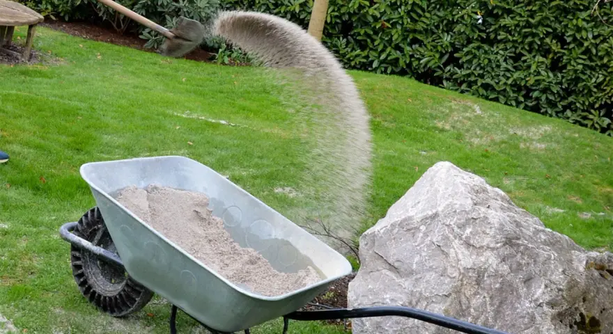 Schubkarre mit Sand, während eine Schaufel Sand auf den Rasen rieselt.