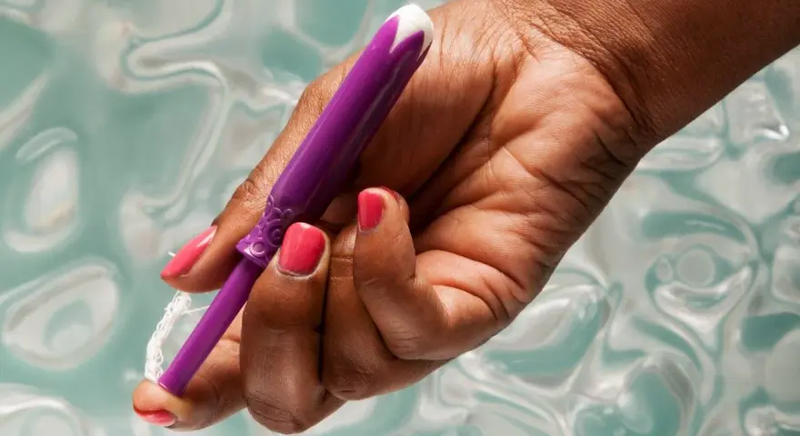 Dłoń z różowym lakierem trzyma fioletowy aplikator tamponu. Pokazano, jak zaaplikować tampon, trzymając go za sznurek.