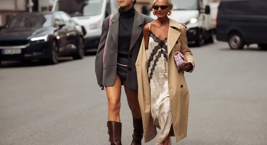 Dos mujeres pasean por la calle. Una lleva un vestido lencero de invierno con un abrigo y la otra un traje gris.