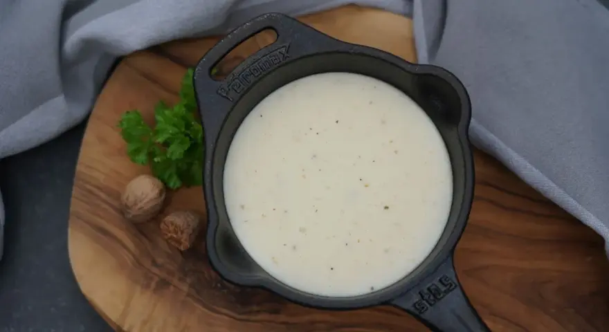 Cremige Béchamelsauce in einer kleinen Gusseisenpfanne, garniert mit Petersilie und Muskatnüssen.