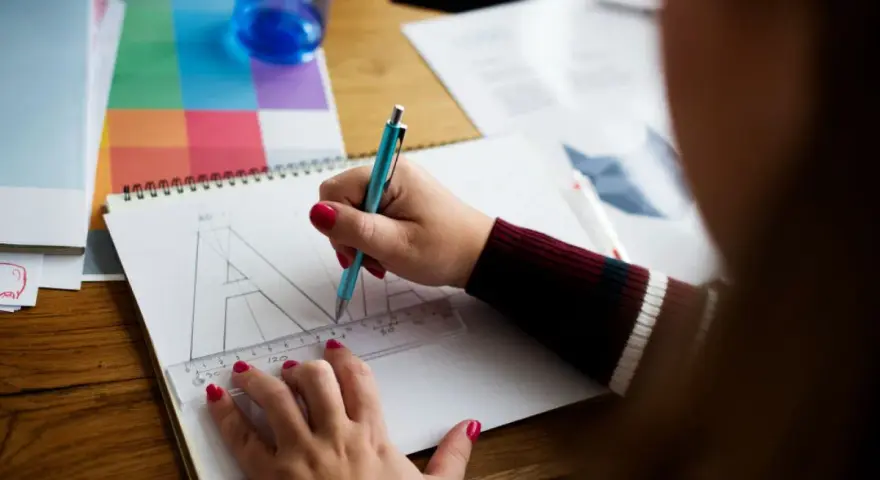 Ilustrator pracuje nad projektem, zamieniając litery na krzywe. Na stole leżą przybory do rysowania i próbnik kolorów.