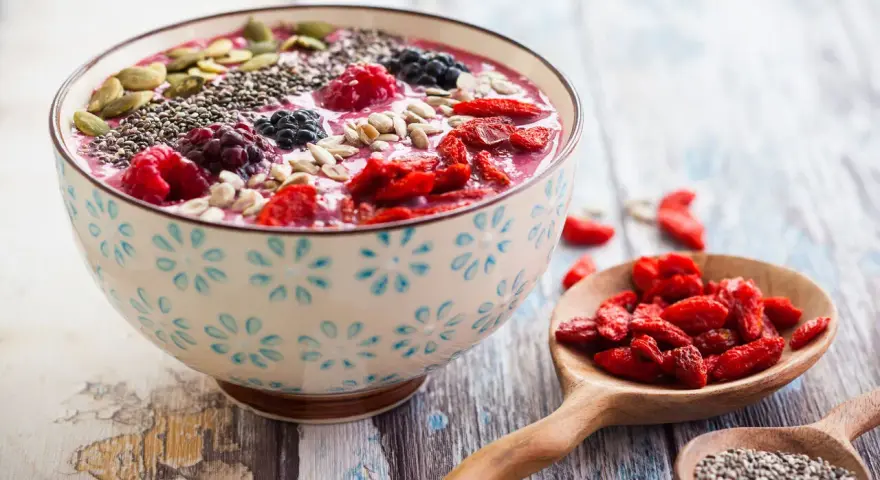 Miseczka smoothie z owocami, nasionami chia, pestkami dyni i słonecznika. Obok łyżki z jagodami goji i nasionami chia. Pyszny sposób, jak jeść nasiona chia.