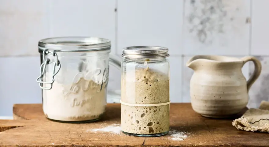 Zwei Gläser mit Sauerteig Weizen, eines mit aktivem, blasigem Sauerteig, das andere mit trockenem Ansatz. Daneben ein kleiner Krug.