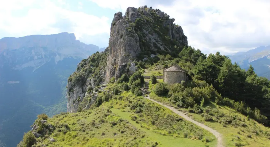 Pequeña ermita de piedra en la ruta de las ermitas, rodeada de vegetación y con imponentes montañas al fondo.