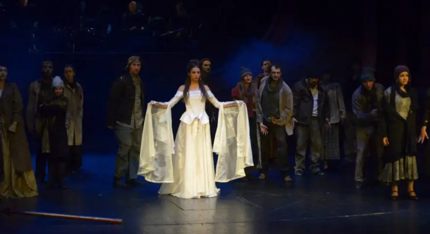 Scena z musicalu "Człowiek z La Manchy". W centrum młoda kobieta w białej sukni, otoczona przez grupę postaci w strojach z epoki.