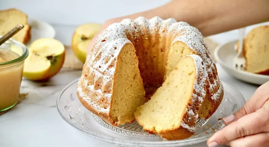Ein saftiger Kuchen mit Apfelmus, bestäubt mit Puderzucker, daneben Äpfel und ein Glas Apfelmus.