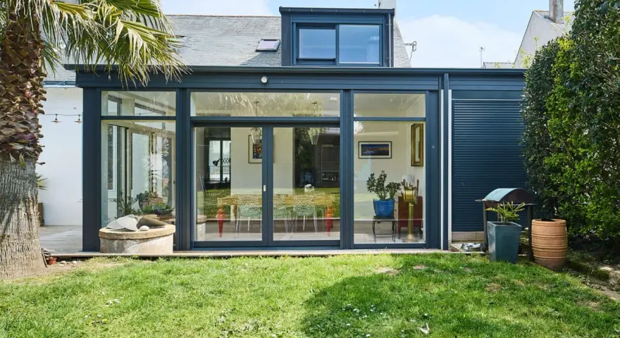 Une extension de maison moderne avec une immense baie vitrée s'ouvrant sur un jardin verdoyant. Une palme se dresse à gauche.