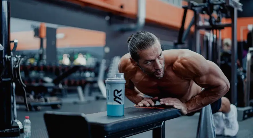 Man gör armhävningar på en bänk i gymmet, en shaker med blå dryck står bredvid. Detta är ett exempel på hur byggs muskler.