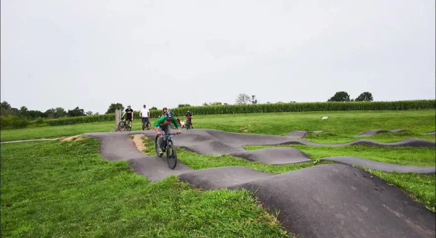A group of people enjoy riding bikes on a winding pump track, with rolling hills and a cornfield in the background.